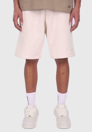 Light beige sweat shorts with a relaxed fit, featuring stitched seams and an embossed brand logo on the lower side. Paired with white socks and sneakers.