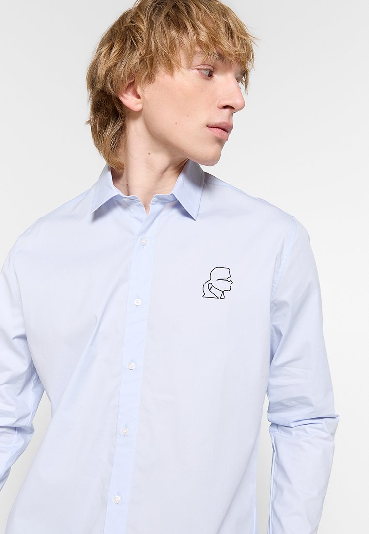 Junge Person mit blonden Haaren, die ein hellblaues, knöpfen Shirt mit einem schwarzen Silhouettenlogo auf der Brust trägt und seitlich schaut.