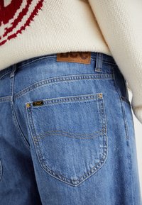 Blauwe denim jeans met een bruine leren label, zichtbare stikdetails en twee achterzakken. Vergezeld door een crèmekleurige gebreide trui.