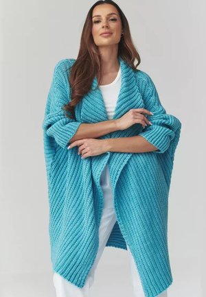 Kobieta ubrana w luźny, gruby turkusowy sweter na drutach, nałożony na biały top i białe spodnie, stojąca na neutralnym tle.