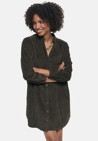 Abito camicia in corduroy verde scuro con bottoni frontali, maniche lunghe e orlo arrotondato. Tessuto testurizzato con dettagli a righe verticali.