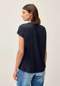 Blusa blu navy a maniche corte con vestibilità ampia, caratterizzata da un dettaglio plissettato sul retro e una superficie liscia, indossata con jeans blu chiaro.