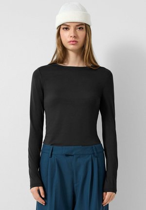 Junge Frau trägt eine weiße Strickmütze, ein schwarzes Langarmshirt und hochtaillierte blaue Hosen, steht vor einem schlichten hellen Hintergrund.