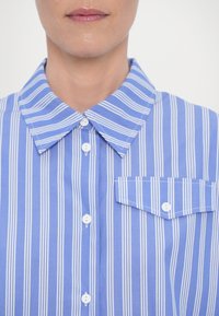 Chemise bleu clair à boutons avec rayures verticales blanches, col pointu et poche plaquée. Le tissu semble lisse et léger.