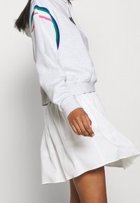 Woman wearing a light gray cropped hoodie with colored stripes on sleeves and a white A-line skirt, side view, against plain background.