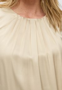 Blouse beige avec un col rond, des plis subtils et une texture douce et lisse. Tissu léger avec un drapé élégant.