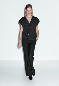 Blusa nera a maniche corte con bottoni e scollatura a V, abbinata a pantaloni neri su misura e scarpe nere, in un ambiente minimalista.