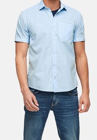 Camicia azzurra a maniche corte in cotone, con un sottile motivo geometrico, taschino frontale e colletto con bottoni. Abbinata a jeans blu.