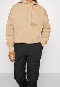 Haut en coton beige avec une poche avant, une capuche à cordon de serrage et un logo imprimé. Associé à un pantalon noir, avec une coupe décontractée.
