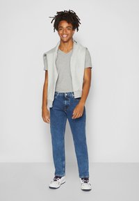 Camiseta gris de cuello en V superpuesta con un cárdigan sin mangas gris claro, jeans de mezclilla azul y zapatillas blancas con acentos rojos y azules.