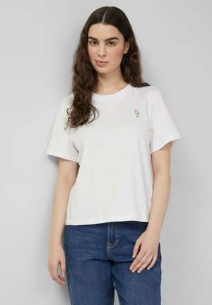 Junge Frau mit langen lockigen Haaren, die ein weißes T-Shirt mit einer kleinen pinken Blumenstickerei und eine blaue Jeans trägt, steht vor einem hellen Hintergrund.