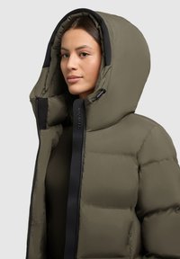 Khaki péřová bunda s velkou kapucí, černým zipem a lemem, s prošívaným vzorem a hladkým povrchem. Logo na táhle zipu.