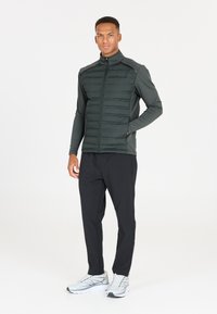 Mørkegrøn puffervest med høj krave og lynlås, kombineret med sorte træningsbukser og grå sneakers med reflekterende detaljer.