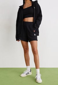 Svart huvtröja, svart kort topp, svarta shorts med vitt logotyp och vita sneakers med grå detaljer. Slät textur, avslappnad design.