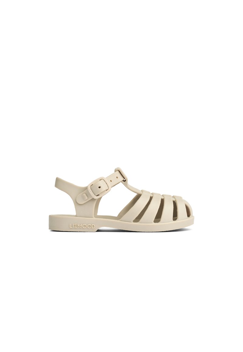 Beige gummisandal med en remdesign, justerbar ankelrem och öppen tå. Har en upphöjd logotyp på sidan och en platt sula.