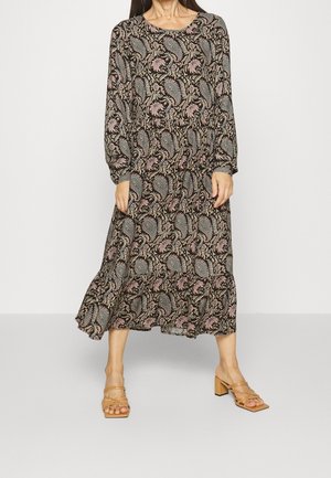 Kobieta w midi sukience z długim rękawem w wzór paisley i kwiatowy, w połączeniu z beżowymi sandałami na słupku, stoi na szarym podłożu.