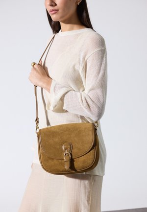 Žena u bijelom džemperu i bež suknji, drži smeđu torbu preko ramena od antilopa s detaljem kopče.