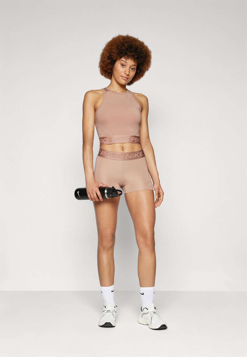 Donna con capelli ricci, in reggiseno sportivo rosa e pantaloni corti, che tiene in mano una bottiglia d'acqua nera, indossando calze bianche Nike e scarpe da ginnastica, in piedi su un pavimento grigio.