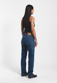 Dark blue denim jeans with a straight cut, featuring a leather patch at the waist and two back pockets. Model shown from the back.