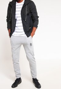 Svart huva jacka med vertikala ränder, vit T-shirt med horisontella ränder, grå sneakersbyxor och svarta sneakers.