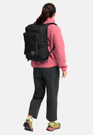 Femme portant un grand sac à dos noir, une veste polaire rose, un pantalon noir et des chaussures de randonnée jaunes, de dos sur un fond blanc uni.