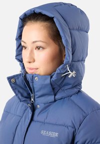 Blå puffjacka med huva, dragkedjestängning och tryckknappsdetaljer. Har ett kviltat mönster och en logotyp på bröstet.