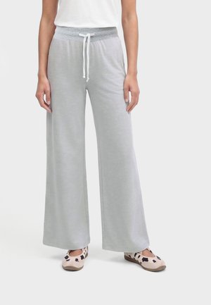 Personne portant un pantalon large gris clair avec taille à cordon et un haut blanc, debout les pieds légèrement écartés et des chaussures à motifs clairs.