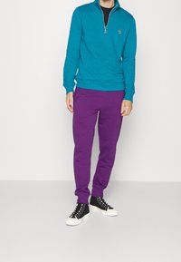 Smaragdgrüner Sweatshirt mit Reißverschluss, violette Jogginghose und schwarze Turnschuhe mit weißen Sohlen. Das Outfit zeigt ein dezentes Logo auf dem Sweatshirt.