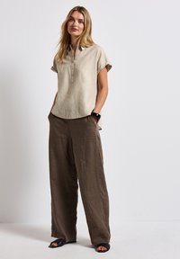 Camicia di lino beige chiaro con colletto e maniche corte arrotolate, abbinata a pantaloni larghi di lino marroni e sandali neri.