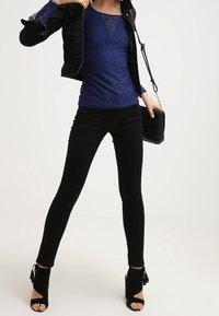 Donna che indossa un top in pizzo blu, giacca nera, pantaloni skinny neri, tacchi aperti neri e tiene una borsa a tracolla nera.