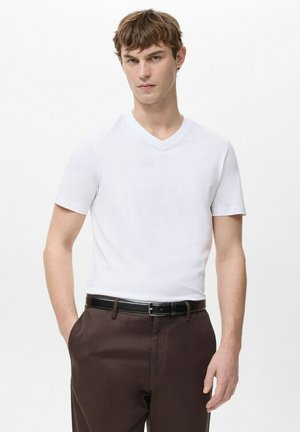 Junger Mann, der ein schlichtes weißes V-Ausschnitt-T-Shirt, braune Hosen und einen schwarzen Gürtel trägt, vor einem weißen Hintergrund steht.