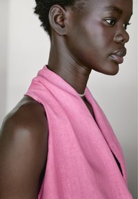 Różowy lniany szal przewieszony wokół szyi, miękka tekstura, z delikatnym połyskiem, podkreślony gładką, ciemną cerą i krótkimi, kręconymi włosami.