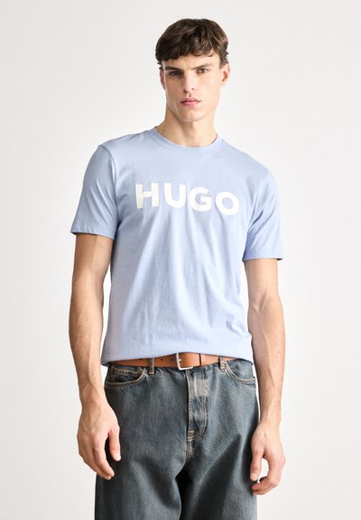 Светлосиня памучна тениска с смел бял принт "HUGO" отпред. Къси ръкави, кръгло деколте, съчетана с тъмносини дънки и кафяв колан.