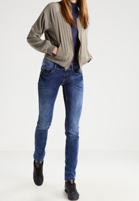 Femme portant une veste grise zippée à l'avant, un col roulé bleu foncé, un jean en denim bleu et des baskets noires, debout les mains dans les poches.