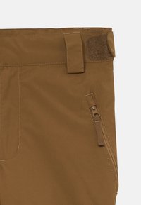 Brune, lette bukser med en teksturert overflate. Den har en sidelomme med glidelås og en borrelåsrem i livet for justering.