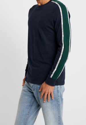 Marineblaues Langarm-T-Shirt mit grünem Streifen und weißem Logoband an den Ärmeln, kombiniert mit hellblauen Jeans. Weicher Baumwollstoff.