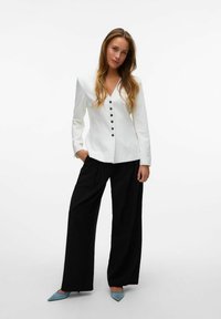 Blazer blanco ajustado con mangas largas y botones negros, combinado con pantalones negros holgados y zapatos planos de punta azul claro.