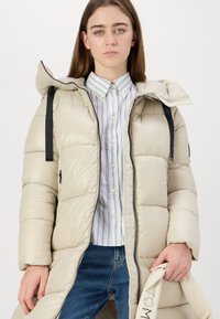 Padded coat in light beige with a hood, featuring black drawstrings, a full zip front, and side pockets. Worn over a striped shirt and jeans.