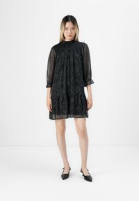 Mulher vestindo um vestido curto preto transparente com padrões de zebra e mangas a três quartos, combinado com sapatos de salto agudo pretos, de pé.