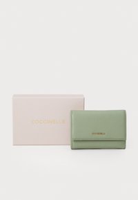 Green leather wallet with a textured finish and gold logo on the front, presented next to a pale pink box featuring the brand name.