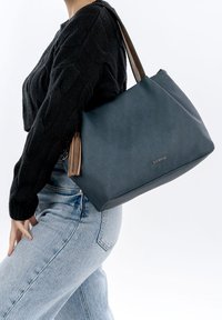 Persoon met een zwarte trui en lichtblauwe spijkerbroek, die een grote blauwe handtas met bruine riemen en kwastje op de schouder draagt.