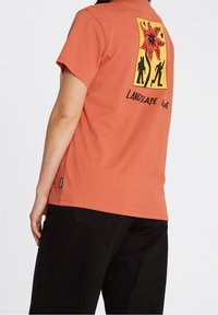 Kortærmet, rustfarvet t-shirt med et grafisk tryk af et rødt blomstermønster og sorte silhuetter på ryggen, med teksten "LANDSCAPE LIE".