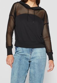 Sweat à capuche noir avec des manches en mesh transparent, un cordon de serrage à la capuche et un corps uni ; associé à un jean bleu clair.