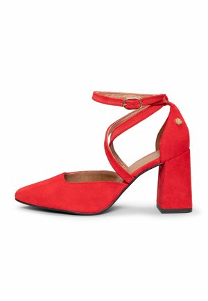 Zapato de tacón grueso de ante rojo con punta puntiaguda y correas cruzadas en el tobillo aseguradas por una pequeña hebilla sobre fondo blanco.