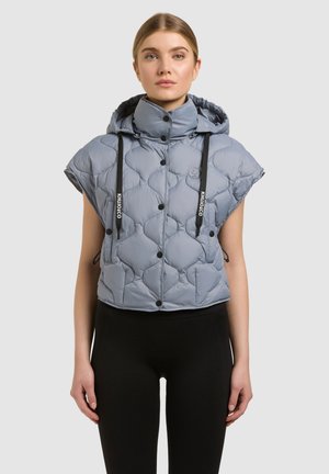 Frau trägt ärmellose, gesteppte graue Kapuzenjacke mit schwarzen Kordeln und schwarzen Knöpfen, kombiniert mit schwarzen Hosen, stehend vor einem grauen Hintergrund.