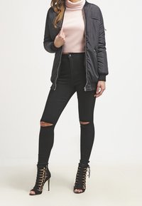 Schwarze Bomberjacke, hellrosa Rollkragenpullover, schwarze zerrissene Skinny-Jeans und schwarze schnürbare High-Heel-Sandalen. Texturiertes Gewebe und schlankes Design.