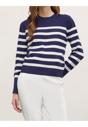 Femme portant un pull à rayures horizontales bleu marine et blanc à manches longues et un pantalon blanc, la main posée sur la hanche.