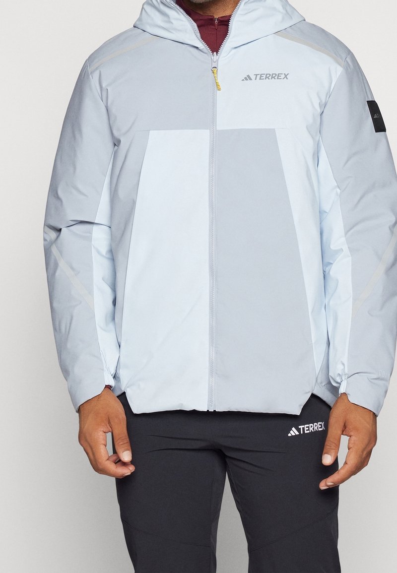 Veste bleu clair avec capuche, fermeture éclair à l'avant, poignets élastiques et accents réfléchissants. Comprend un logo sur la poitrine et un panneau avant contrasté.