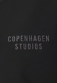Crna tkanina s izvezenim tekstom "Copenhagen Studios" u svijetlo sivoj boji, koja prikazuje glatku teksturu i jednostavan, minimalistički dizajn.