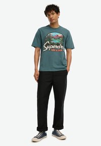 Camiseta de algodón color verde azulado con un gráfico colorido de montaña y el texto "Superdry Take a Trip". Combinada con pantalones negros y zapatillas azules.